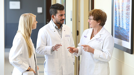 A group of three clinicians wearing white coats talk with their hands in a hallway.